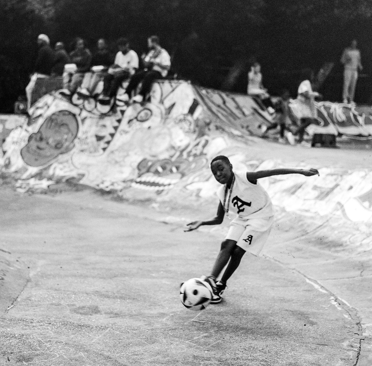 Boy dribbling a ball through a graffiti skate bowl, black and white