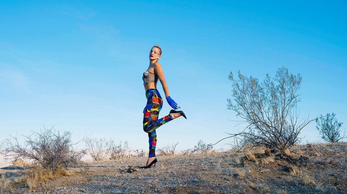 Model in patterned mesh and blue gloves kicking a heel up against a clear blue sky
