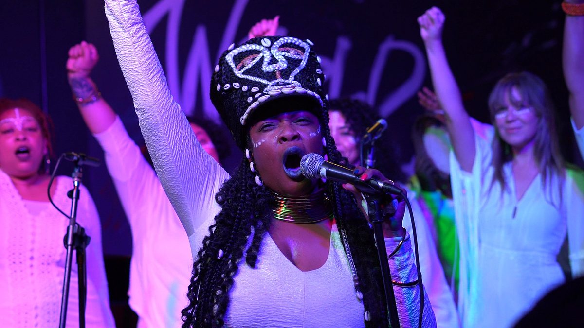 Singer in a cowrie-shell crown, fist raised, with backing singers behind