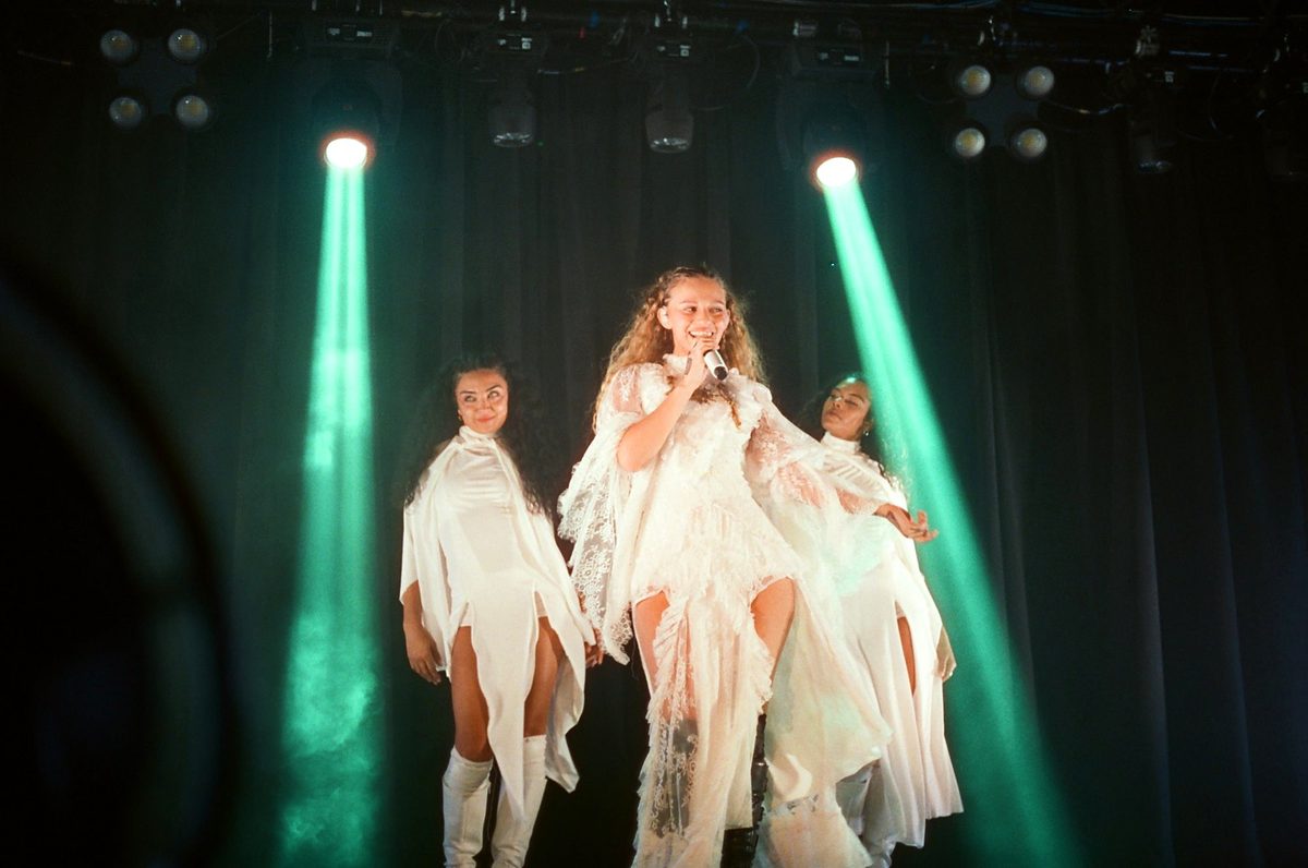 Singer in white flanked by dancers under green beams