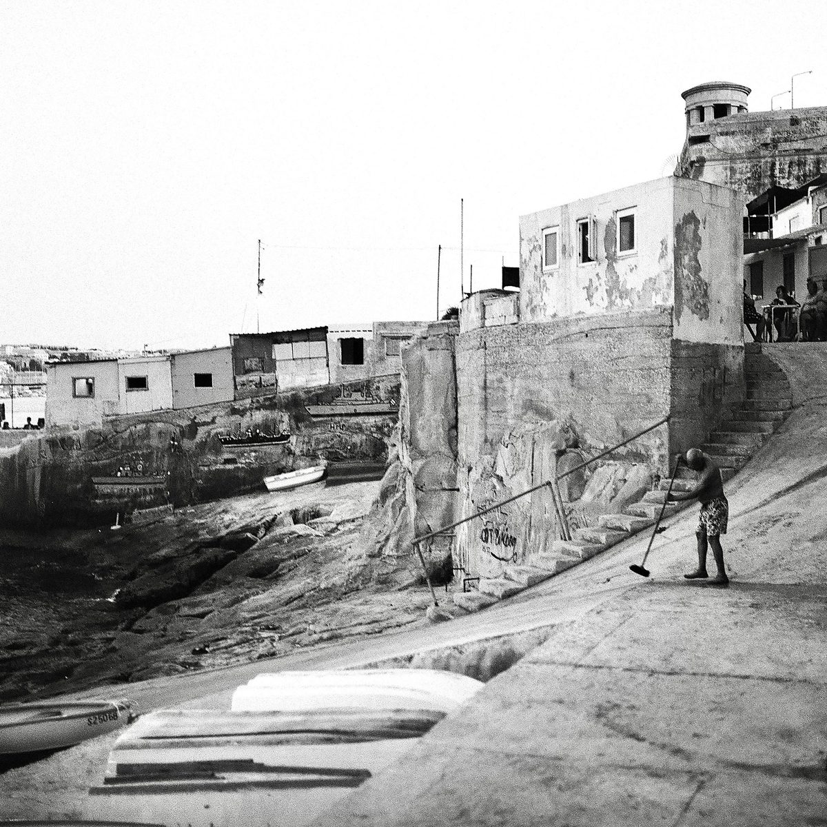 Man sweeping a waterfront slipway below a fortress, black and white